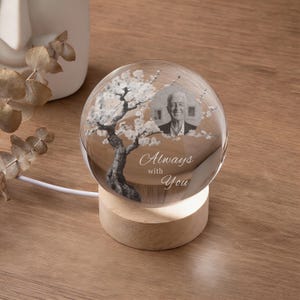 May include: A clear glass sphere with a photo of a person and a tree design, resting on a wooden base. The sphere has the words "Always with You" etched on it. The base is illuminated by a soft light.