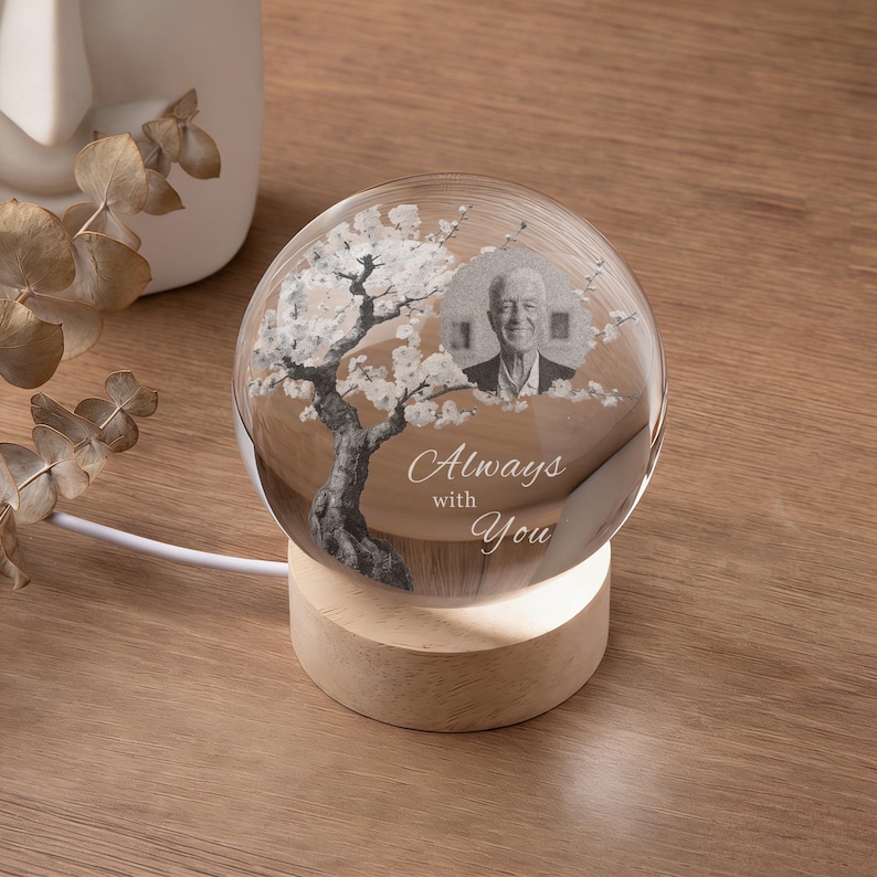 May include: A clear glass sphere with a photo of a person and a tree design, resting on a wooden base. The sphere has the words "Always with You" etched on it. The base is illuminated by a soft light.