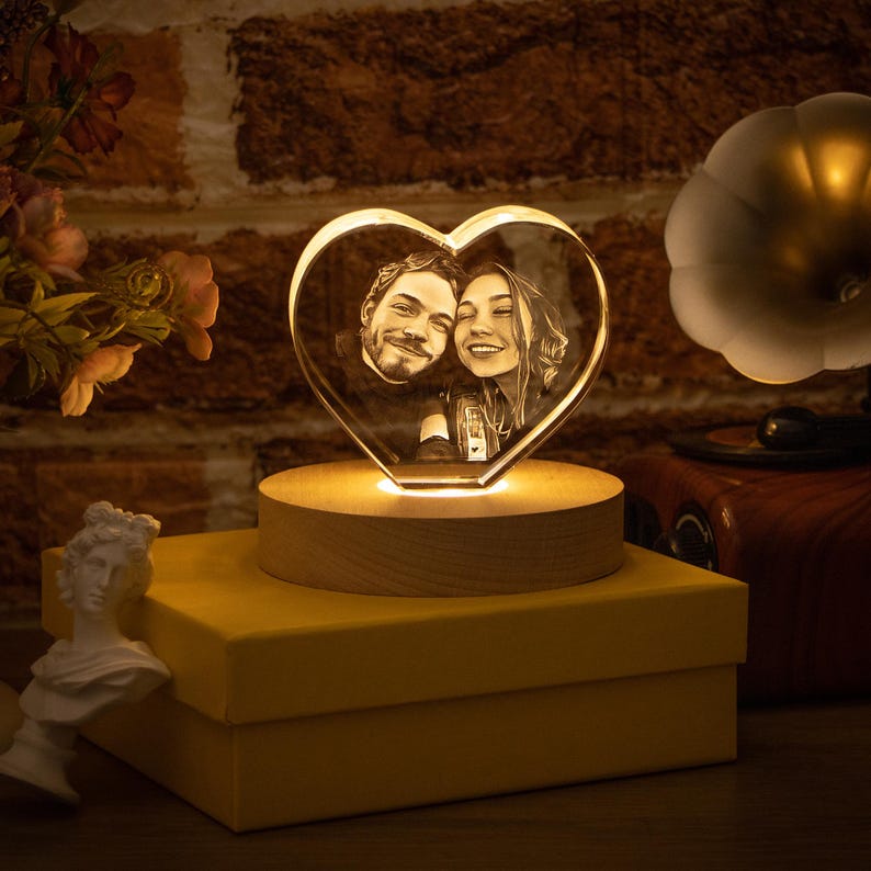 May include: A heart-shaped crystal photo, lit from within, displaying a couple's portrait. It rests on a wooden base, placed on a yellow box. The background features a brick wall and a gramophone.