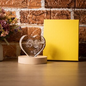 May include: A heart-shaped crystal photo featuring a couple's image, mounted on a wooden base. A yellow box is positioned behind the crystal, with a brick wall backdrop and flowers on the left.