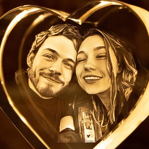May include: A heart-shaped, illuminated glass art piece featuring a smiling couple. The faces are etched within the glass, creating a 3D effect. The warm, golden light enhances the romantic theme, perfect for a personalized gift.