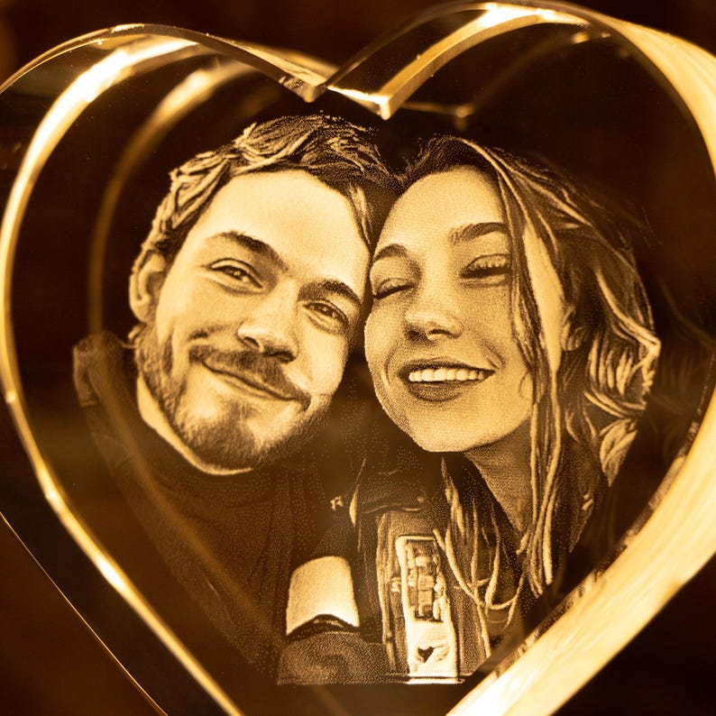 May include: A heart-shaped, illuminated glass art piece featuring a smiling couple. The faces are etched within the glass, creating a 3D effect. The warm, golden light enhances the romantic theme, perfect for a personalized gift.