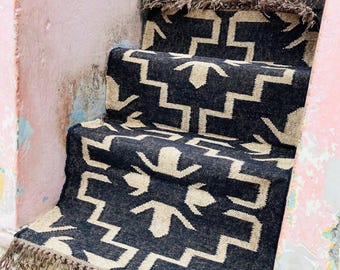 May include: A black and beige patterned stair runner with a geometric design and fringed edges. The runner is placed on weathered concrete steps with peeling pink paint.