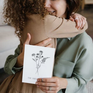 Puede incluir: Una mujer y una ni&ntilde;a se abrazan mientras sostienen una tarjeta con un dibujo en blanco y negro de flores y el texto "THE SMITHS".