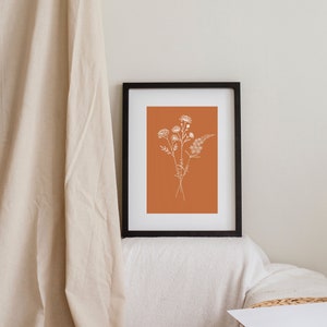 Puede incluir: Una impresi&oacute;n enmarcada con un dibujo minimalista de un ramo de flores en blanco sobre un fondo naranja quemado.