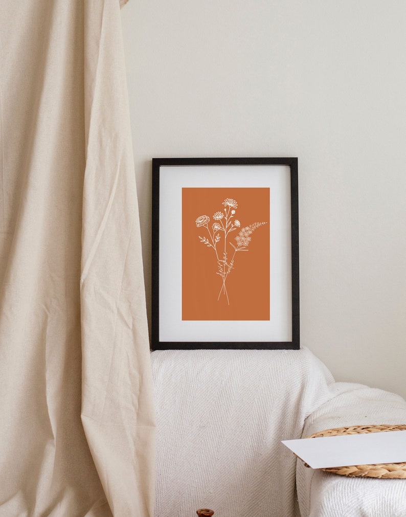 Puede incluir: Una impresi&oacute;n enmarcada con un dibujo minimalista de un ramo de flores en blanco sobre un fondo naranja quemado.