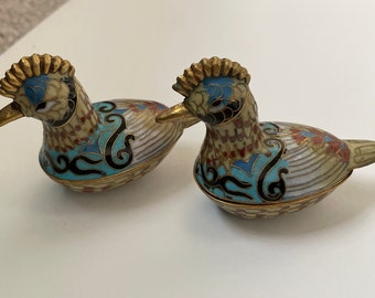 Op de afbeelding: Twee kleine, ingewikkeld gedetailleerde cloisonné-emaille vogelbeeldjes. De vogels zitten met gevouwen vleugels. De vogels zijn versierd met verschillende kleuren, waaronder blauw, groen, rood en goud.