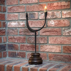 May include: A black metal candle holder with a wooden base. The candle holder is shaped like a U and has a single candle holder on the top. The candle holder is sitting on a brick wall.