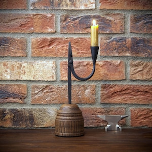 May include: A handcrafted candle holder with a lit yellow candle. The candle holder has a dark metal arm and a wooden base. The background is a brick wall. A small metal anvil is on the wooden surface.