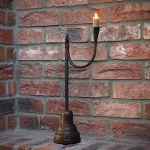 May include: A black metal candle holder with a single candle lit. The holder is attached to a tall, thin, black metal base. The candle holder is in front of a brick wall.