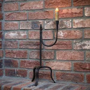 May include: A black metal candle holder with a single arm and a candle lit inside. The holder is standing on a brick surface.