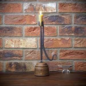 May include: A lit candle in a handcrafted metal and wood candle holder. The candle is yellow. The holder has a dark metal arm and a wooden base. The background is a brick wall. A small metal anvil is on the right.