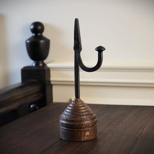May include: A black metal hook with a wooden base. The hook is attached to a round wooden base with a fluted design.
