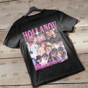 May include: Black t-shirt with a pink graphic that says "HOLLANOV" and "Heated Rivalry." The graphic features a collage of images of people. The shirt is hanging on a black hanger.
