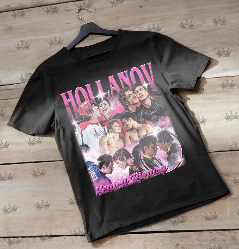 May include: Black t-shirt with a pink graphic that says "HOLLANOV" and "Heated Rivalry." The graphic features a collage of images of people. The shirt is hanging on a black hanger.