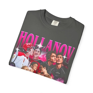 May include: Dark gray t-shirt featuring a group of young men and the word "HOLLANOV" in pink, retro-style lettering. The shirt has a crew neck and a tag in the upper right corner.