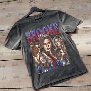 May include: Dark gray t-shirt featuring a graphic of Brooke Davis with the text "Brooke Davis" in a gradient red and blue font. The shirt displays multiple images of the actress. The shirt is hanging on a black hanger.