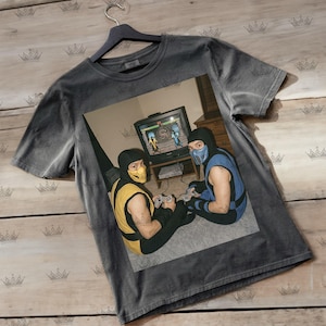 May include: Dark gray t-shirt featuring a graphic of two people dressed as Mortal Kombat characters playing a video game on an old television. The characters are Scorpion and Sub-Zero. The shirt is hanging on a black hanger.