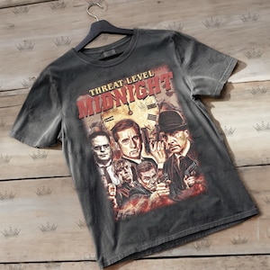 May include: Dark gray t-shirt featuring a graphic with the text "Threat Level Midnight" in red. The graphic includes images of several people with guns and a clock face. The shirt is hanging on a black hanger.