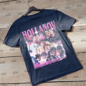 May include: Navy blue t-shirt featuring a collage of images with the text "HOLLANOV" in pink, and "Heated Rivalry" in pink. The images include people in various poses, some shirtless, and some in hockey gear.