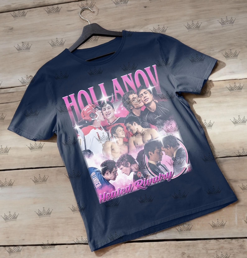 May include: Navy blue t-shirt featuring a collage of images with the text "HOLLANOV" in pink, and "Heated Rivalry" in pink. The images include people in various poses, some shirtless, and some in hockey gear.