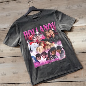Könnte beinhalten: Dunkelgraues T-Shirt mit einem rosa Grafikdesign. Das Design enthält das Wort "HOLLANOV" und Bilder von Personen. Die Worte "Heated Rivalry" sind ebenfalls in Rosa.