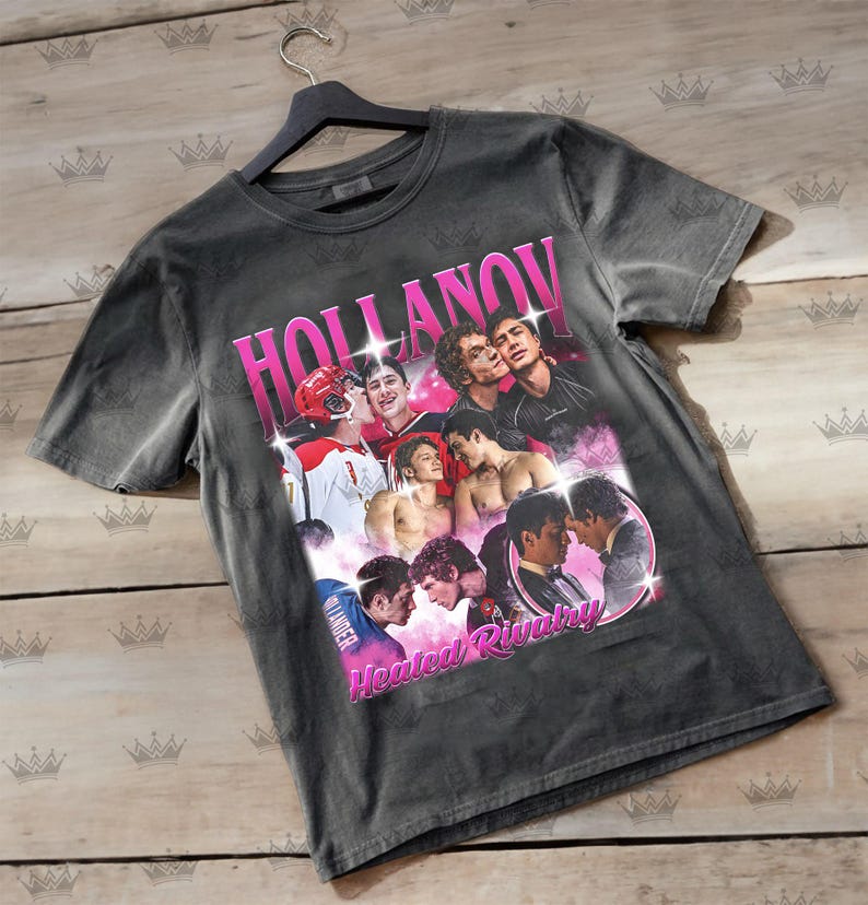 May include: Dark gray t-shirt featuring a graphic design with the word "HOLLANOV" in pink, along with images of several people. The words "Heated Rivalry" are also in pink.