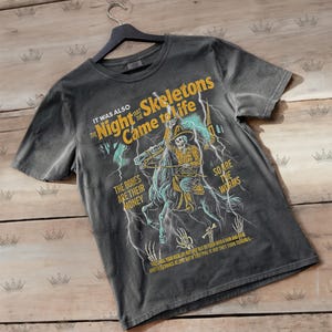 Könnte beinhalten: Ein anthrazitfarbenes Kurzarm-T-Shirt mit einer Grafik eines Skelett-Cowboys auf einem Pferd inmitten von Blitzen. Der Text lautet: IT WAS ALSO THE NIGHT THAT THE SKELETONS CAME TO LIFE, THE BONES ARE THEIR MONEY, SO ARE THE WORMS.