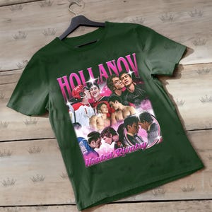 May include: Dark green t-shirt featuring a collage of images with the text "HOLLANOV" in pink, and "Heated Rivalry" in pink. The images depict various people in different poses. The shirt is hanging on a black hanger.