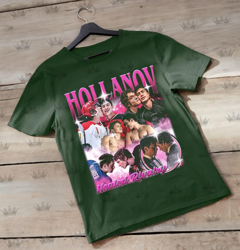 May include: Dark green t-shirt featuring a collage of images with the text "HOLLANOV" in pink, and "Heated Rivalry" in pink. The images depict various people in different poses. The shirt is hanging on a black hanger.