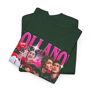 May include: Dark green t-shirt featuring a graphic of a group of young men, with the word "OLLANO" in large pink letters. One man is wearing a red hockey helmet. The t-shirt is folded.