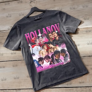 May include: Dark gray t-shirt featuring a collage of images with the text "HOLLANOV" in pink, and "Heated Rivalry" in pink. The images include people in hockey uniforms and other casual wear.