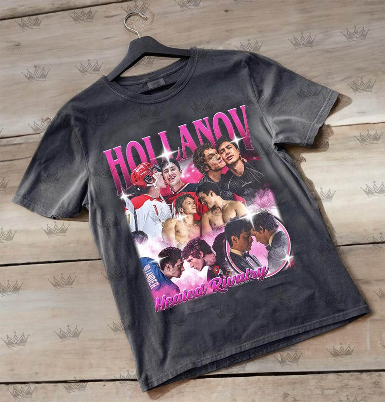 May include: Dark gray t-shirt featuring a collage of images with the text "HOLLANOV" in pink, and "Heated Rivalry" in pink. The images include people in hockey uniforms and other casual wear.