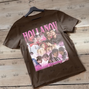 May include: A brown t-shirt featuring a collage of images with the text "HOLLANOV" in pink, and "Heated Rivalry" in pink. The shirt is hanging on a black hanger against a light wood background.