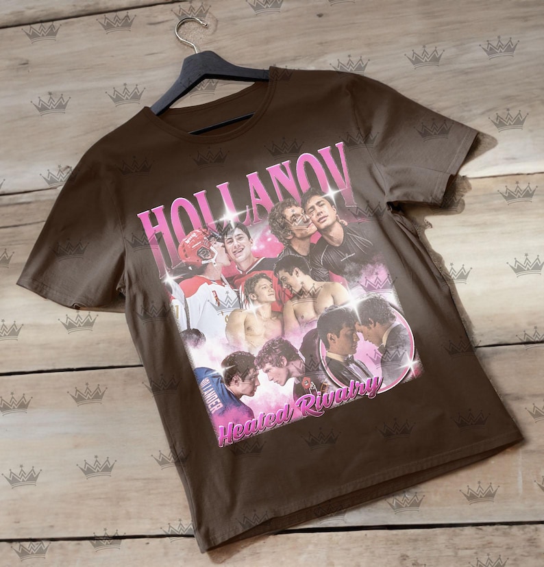 May include: A brown t-shirt featuring a collage of images with the text "HOLLANOV" in pink, and "Heated Rivalry" in pink. The shirt is hanging on a black hanger against a light wood background.