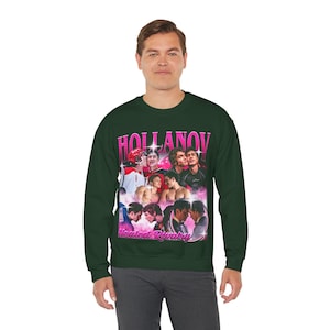 May include: Dark green sweatshirt featuring a graphic design with the word "HOLLANOV" in pink, along with images of people. Below the main text is the phrase "Heated Rivalry" in pink. The sweatshirt is a crew neck style.