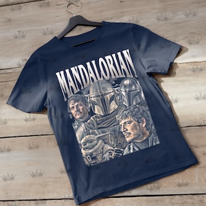 Pu&ograve; includere: T-shirt blu navy con la scritta "MANDALORIAN" in bianco. La maglietta presenta una grafica dei personaggi della serie, tra cui il Mandaloriano e Grogu. La maglietta &egrave; appesa a una gruccia nera.