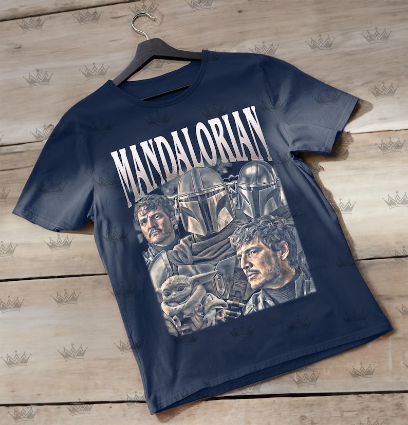 Pu&ograve; includere: T-shirt blu navy con la scritta "MANDALORIAN" in bianco. La maglietta presenta una grafica dei personaggi della serie, tra cui il Mandaloriano e Grogu. La maglietta &egrave; appesa a una gruccia nera.