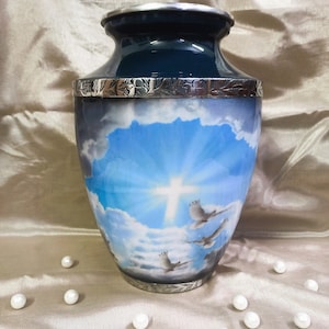 May include: A blue and silver cremation urn with a white cross and birds flying in a blue sky. The urn has a silver trim and a floral design.