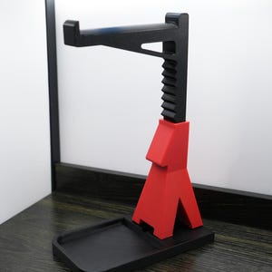 May include: A black and red headphone stand. The stand has a rectangular base with a small tray, a red support structure, and a black adjustable arm to hold headphones. The stand is made of plastic.