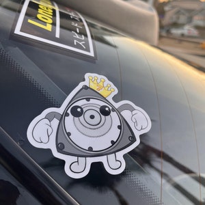 Könnte beinhalten: Ein weißer Aufkleber mit einer Cartoon-Illustration eines grauen Autoteils, das eine goldene Krone trägt. Das Autoteil hat große Augen und lächelt. Der Aufkleber befindet sich auf einer schwarzen Autoscheibe.