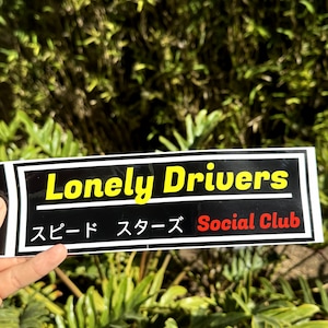 May include: A black and white sticker with the text "Lonely Drivers" in yellow and "Social Club" in red. The text "スピード スターズ" is in black and white.