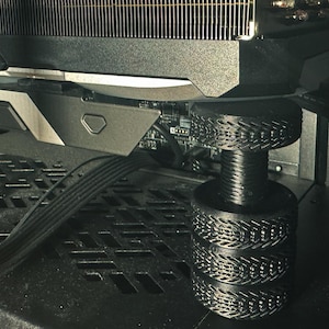 May include: Close-up of a black computer component with a patterned support structure. The component is part of a computer's internal hardware, likely a graphics card or cooling system. The image is taken from a low angle, highlighting the intricate design.