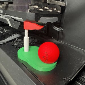 May include: A 3D-printed green golf putting green base with a white pole and a red flag, featuring a bright red Callaway golf ball with the number 3 printed on it. The object is positioned beneath a black computer graphics card component.