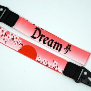 May include: Two pink and white lanyard keychains with black clips. One keychain has the word "Dream" and the Japanese word for dream written in black. The other keychain has a red sun and white cherry blossoms.