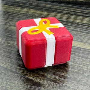 May include: A small, cube-shaped object resembling a gift box, featuring a red base with a white cross-shaped ribbon design and a yellow bow on top. The surface of the object shows visible horizontal layers from a 3D printing process.