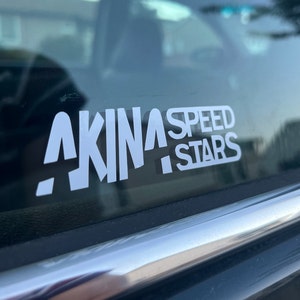 Op de afbeelding: Witte vinyl sticker met de tekst "AKINA SPEED STARS" op een zwart autovenster.