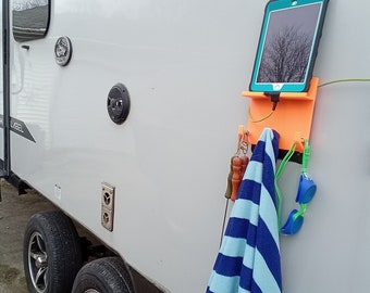May include: An orange tablet holder with a teal tablet, a blue and white striped towel, and swim goggles hanging on the side of a white RV. The RV has black tires and a black speaker.