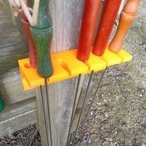 May include: Orange plastic wall mount with five slots for holding metal skewers. The skewers are inserted into the slots and hang down. The wall mount is attached to a wooden post.