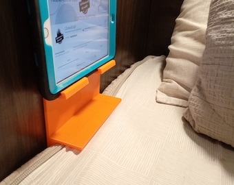 Puede incluir: Un soporte naranja para tableta con estante montado en la pared. Una tableta turquesa está asegurada en el soporte, mostrando un sitio web con el texto "ALWAYS HANDCRAFTED" y un logotipo. El soporte está junto a una cama con una colcha estampada.